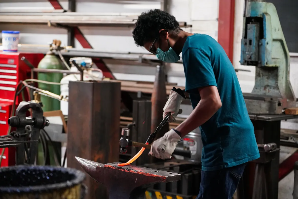 Man wearing a mask hammering molten metal in workshop.